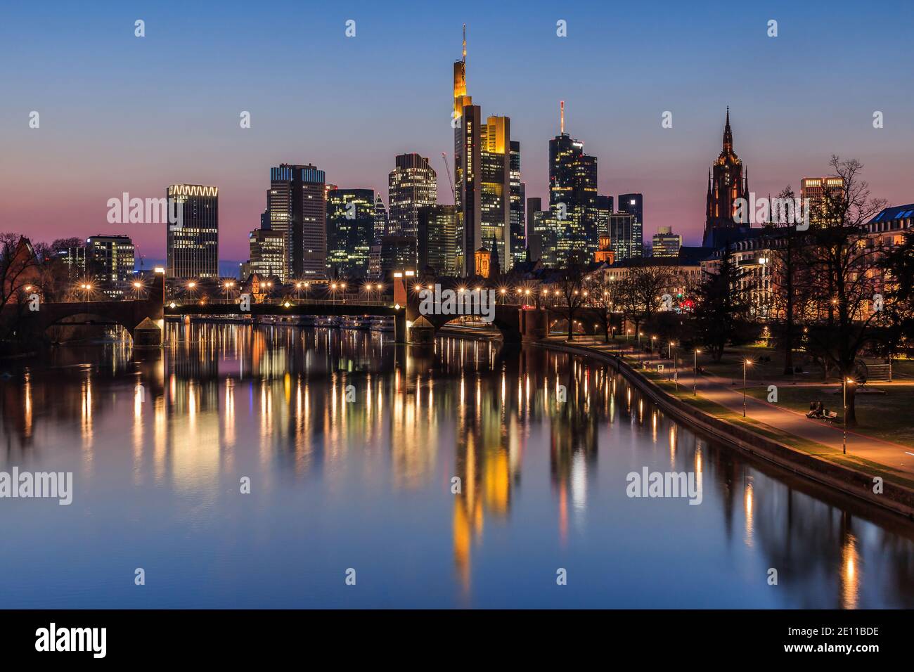 Frankfurter Skyline am Abend zur blauen Stunde. Main mit Spiegelungen von Hochhäusern. Beleuchtete Geschäftshäuser mit einer Brücke Stockfoto