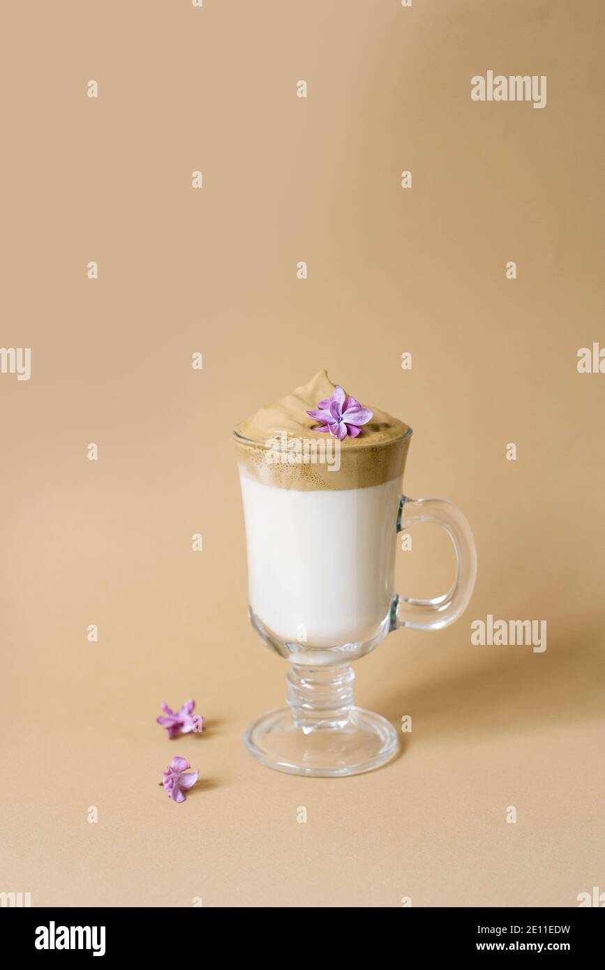 Schöner Dalgon-Kaffeedrink mit Schaum in einer transparenten Tasse und lila Blüten auf beigem Hintergrund. Speicherplatz kopieren Stockfoto