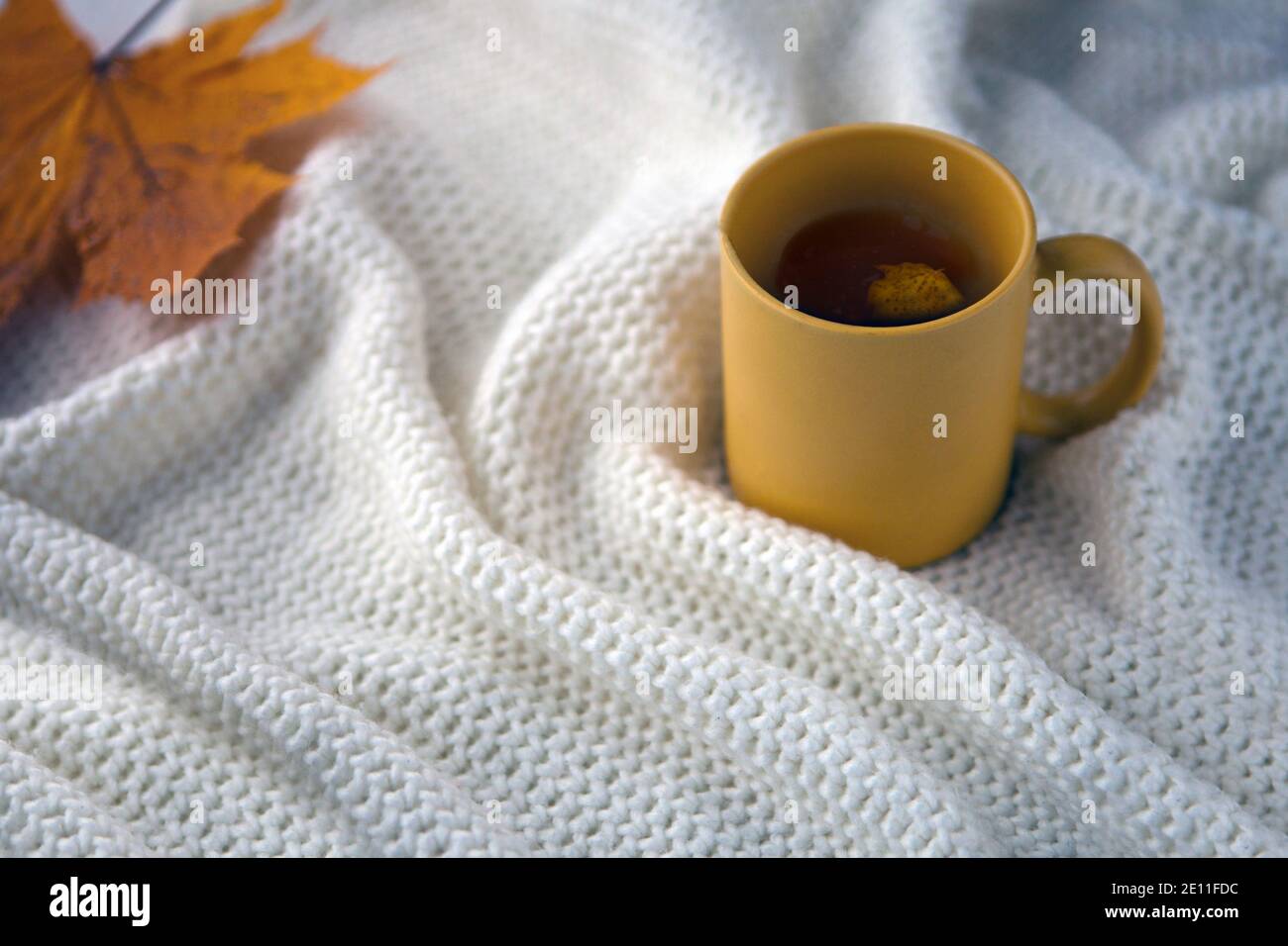 Gelbe Tee-Tasse mit weißem Schal Hintergrund, Keramik-Tasse mit Herbstblatt, weiße Decke mit heißem Getränk Stockfoto