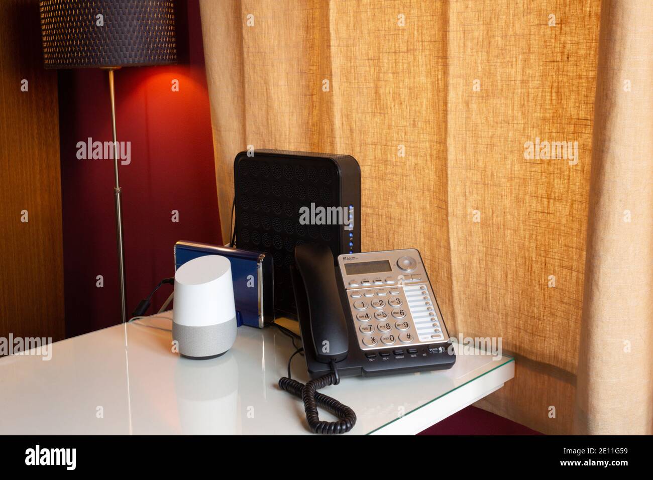 Home Office Schreibtisch mit einem sprachaktivierten, virtuellen Smart Assistant, einem Festnetztelefon und zwei Routern auf einem weißen Schreibtisch mit einer Lampe beleuchtet Stockfoto