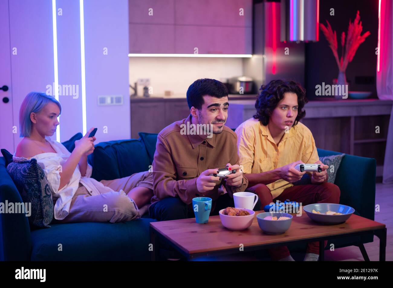 Zwei männliche Freunde aufgeregt, Videospiele zu Hause zu spielen, während ein Mädchen mit ihnen auf einer Couch und mit Handy-Smartphone sitzen. Stockfoto