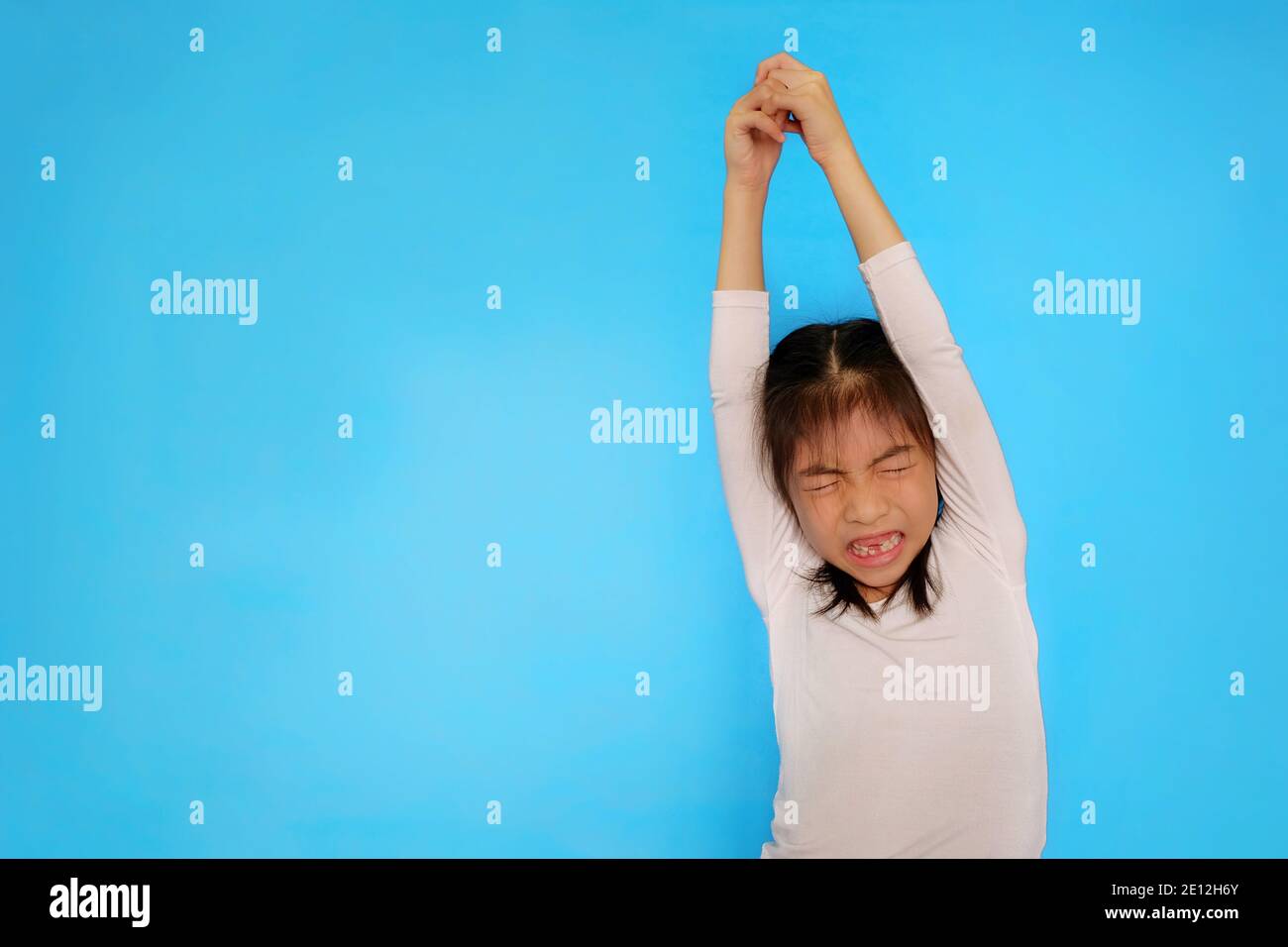Ein süßes junges asiatisches Mädchen mit einem weißen Hemd dehnt sich mit ihren Armen nach oben und ihren Händen zusammen, bevor sie eine Übung beginnt. Einfarbiges hellblaues Backgro Stockfoto