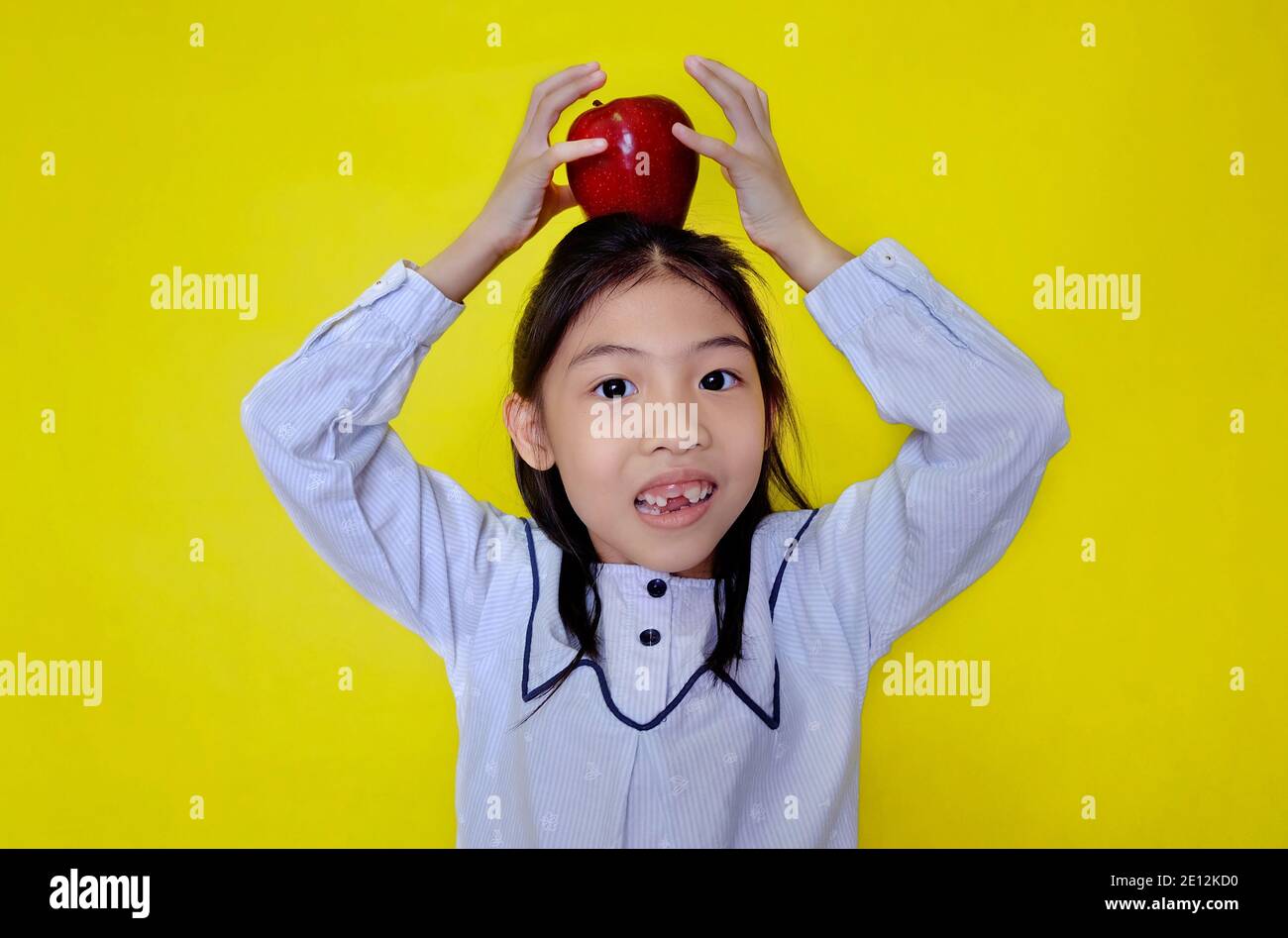 Ein süßes junges asiatisches Mädchen, das versucht, einen roten Apfel oben auf ihrem Kopf zu balancieren, ihn mit ihren Händen zu verkrampfen, bevor es loslässt. Hellgelber Hintergrund. Stockfoto