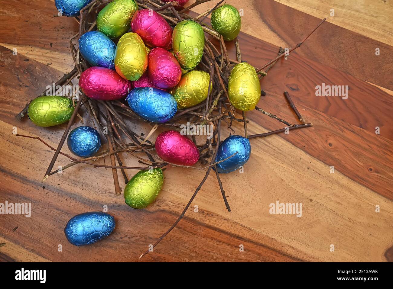 In Folie gewickelte bunte ostereier in rosa, grün, blau und gelb in einem natürlichen Nest aus Stöcken und Zweigen, vor einem braunen Holzhintergrund. Stockfoto