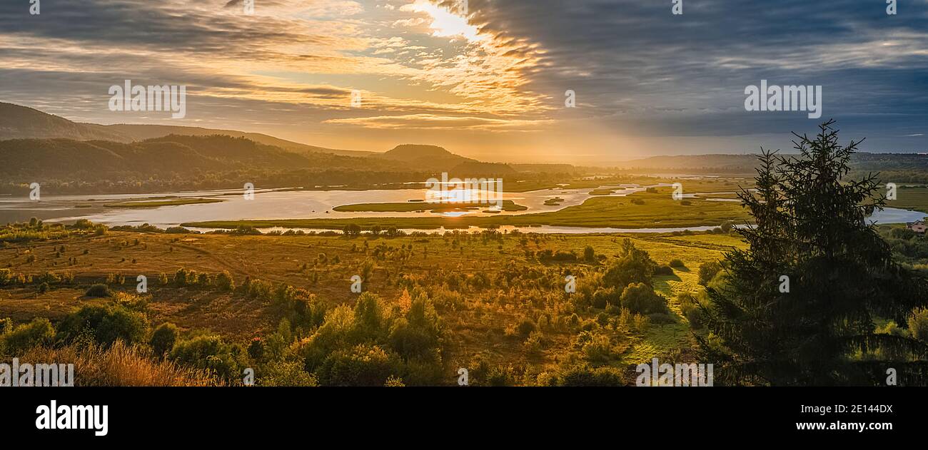 Wunderschöne Landschaft mit Bergen und Hügeln, Fluss und Bäumen, die von den Strahlen der aufgehenden Sonne durch die Wolken am Himmel beleuchtet werden. Bildschirmschoner, l Stockfoto