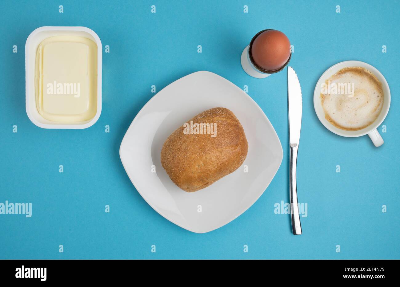 Direkt über Ansicht von Brötchen auf Teller, Butter, gekochtem Ei, Messer und Tasse Kaffee auf blauem Hintergrund, Frühstückskonzept Stockfoto