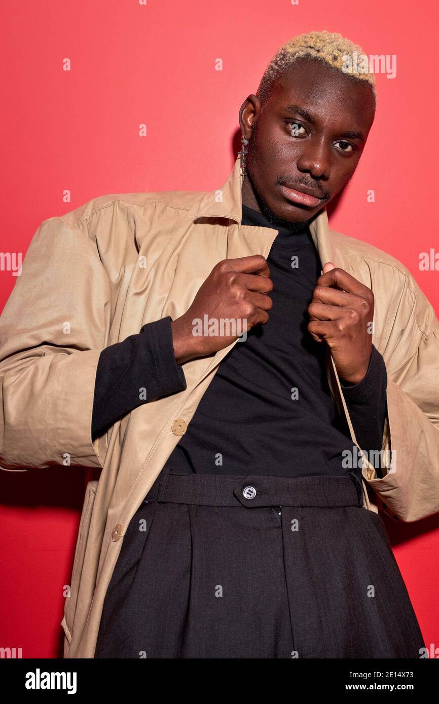 afrikanischer Mann in beigefarbenem Graben und schwarzem tragen innen des Mantels, posiert vor der Kamera isoliert über rotem Studiohintergrund Stockfoto