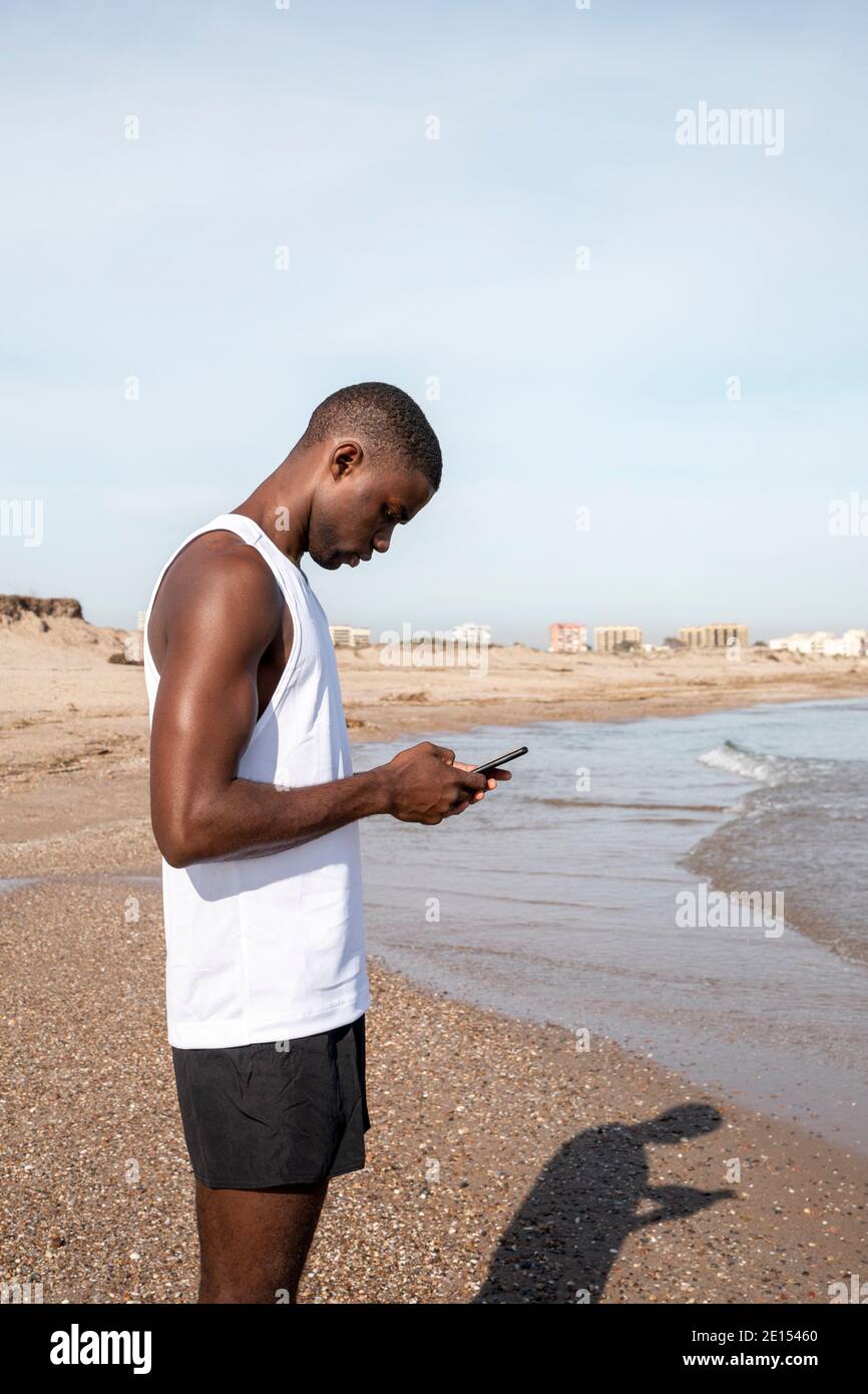 Vertikale Aufnahme eines jungen schwarzen Sportlers vor dem Meer, der sein Handy benutzt. Konzept der Konnektivität. Stockfoto