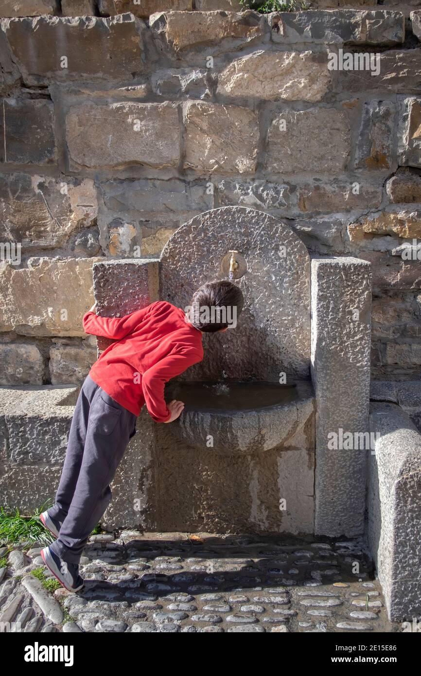 Kind auf dem Rücken trinkt Wasser auf Zehenspitzen in einem Steinbrunnen in Ainsa, Huesca Spanien, vertikal Stockfoto