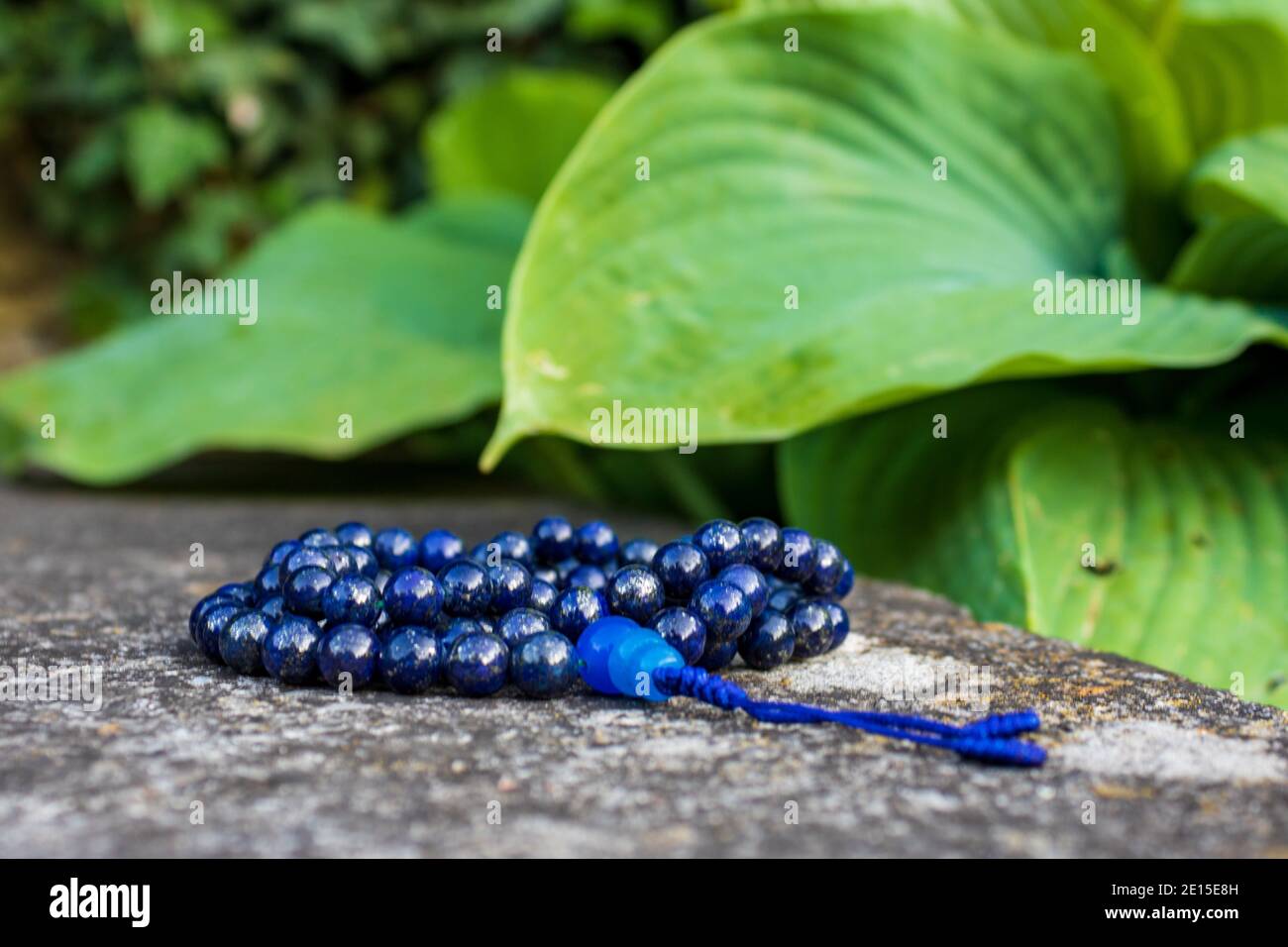 Blaue mala Perlen auf Steinwand mit grünen Blättern im Hintergrund, selektiver Fokus. Yoga und Meditation Zubehör. Stockfoto