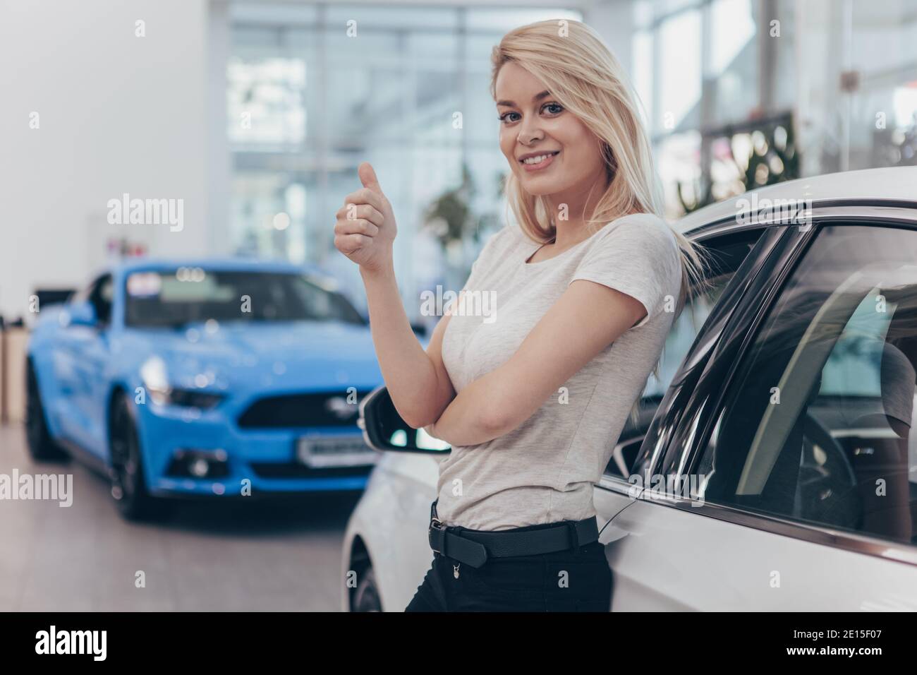 Aufgeregt junge schöne weibliche Fahrerin posiert in der Nähe ihres neu gekauften Autos im Autohaus, zeigt Daumen nach oben, Copy Space. Wunderschöne Frau beim Kauf Stockfoto