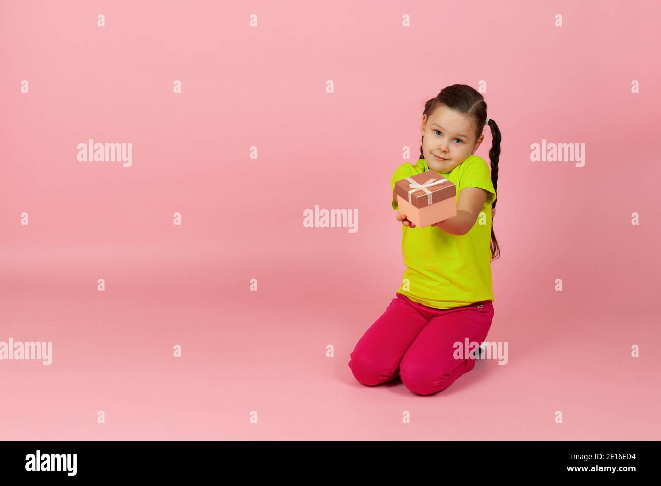 Mock up Mädchen mit Zöpfen in einem hellgrünen T-Shirt und roten Hosen reicht uns eine Geschenkbox auf dem Boden sitzen , isoliert auf einem rosa Hintergrund Stockfoto