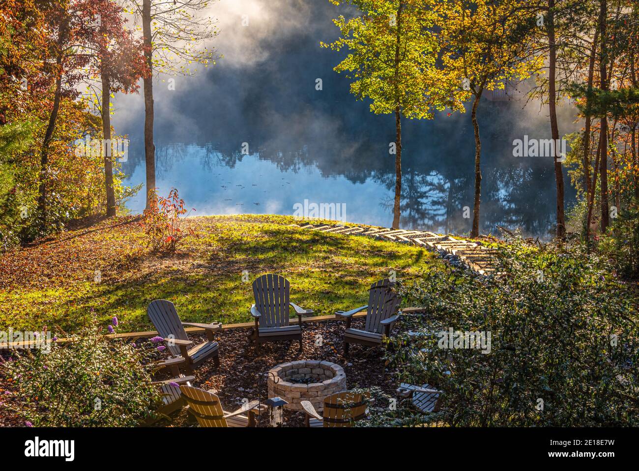 Nebel steigt von einem Bergsee auf, während die aufgehende Sonne Herbstlaub um ein gehobenes Blockhaus-See-Haus in den Nord-Georgia-Bergen erhellt. Stockfoto