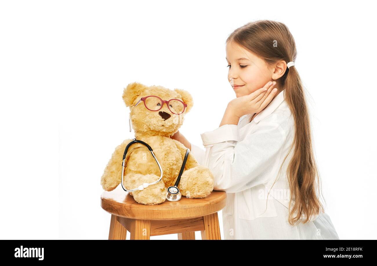 Kaukasisches Mädchen, das einen Arztmantel trägt, spielt mit ihrem Spielzeugbären-Patienten. Isolierter weißer Hintergrund Stockfoto