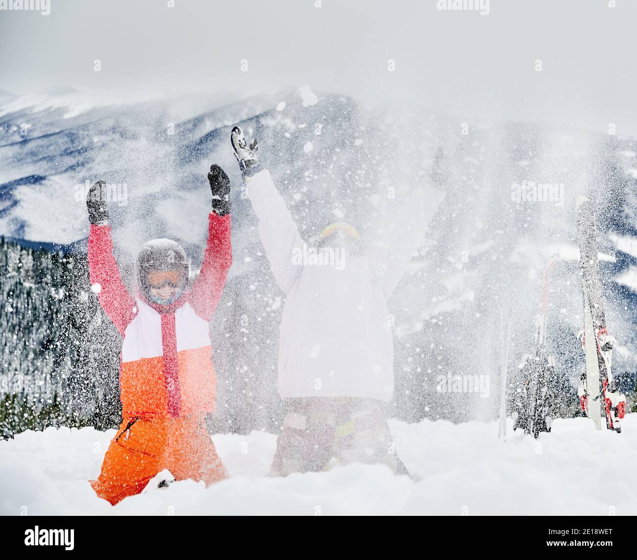 Zwei Skifahrer in Skianzügen und Helmen werfen frischen Pulverschnee hoch in die Luft, Spaß im Skigebiet mit Bergen im Hintergrund. Konzept der Wintersport-Aktivitäten, Spaß und Beziehungen. Stockfoto