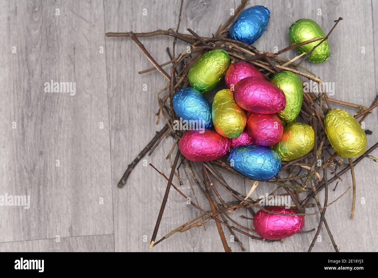 In Folie gewickelte mehrfarbige ostereier in rosa, grün, blau und gelb in einem natürlichen Nest, aus Stäben und Zweigen, auf einem grauen weißen Holzhintergrund. Stockfoto
