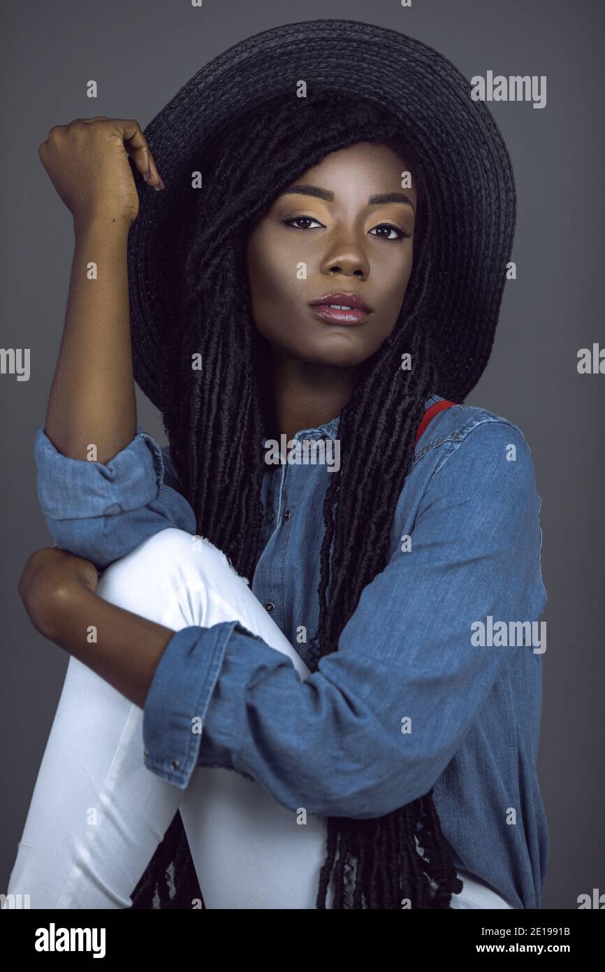 Porträt einer schönen jungen schwarzen Frau mit langen Dreadlocks & schönes Make-up posiert von selbst tragen bunten Schmuck, Sommerhut, Denim-Shirt Stockfoto