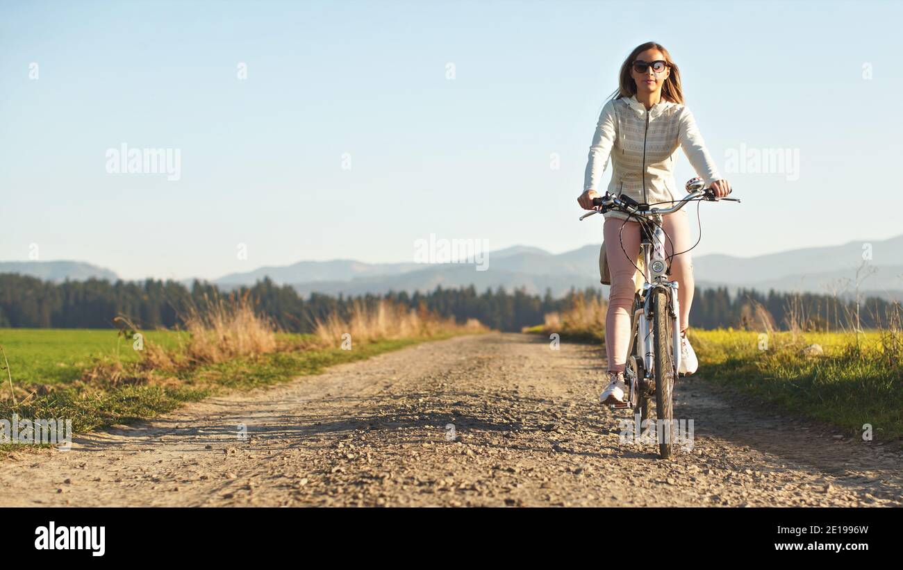 Junge Frau in Jacke und Sonnenbrille fährt mit dem Fahrrad über die Landstraße, Nachmittagssonne scheint auf verschwommenem Hintergrund, Platz für Text links Stockfoto