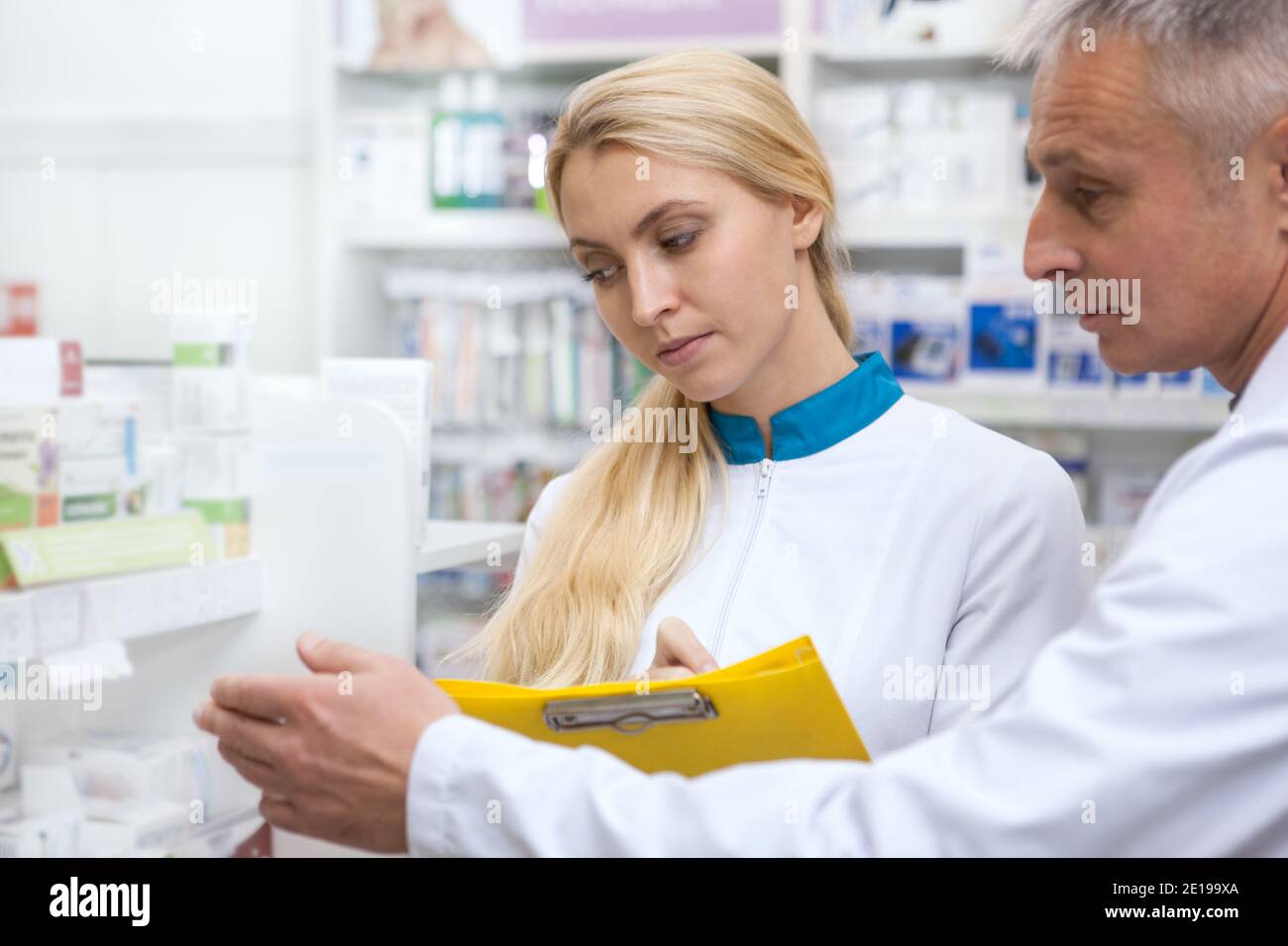 Schöne junge Apothekerin, die in der Drogerie mit ihrer älteren Kollegin arbeitet und Produkte in den Regalen untersucht. Reif männlich Chemiker im Gespräch mit seine y Stockfoto