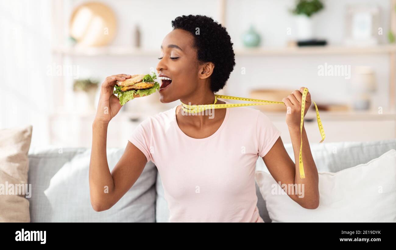 Afroamerikanische Frau, die Cheeseburger essen will, zieht sich zu Hause mit Maßband weg, Panorama Stockfoto