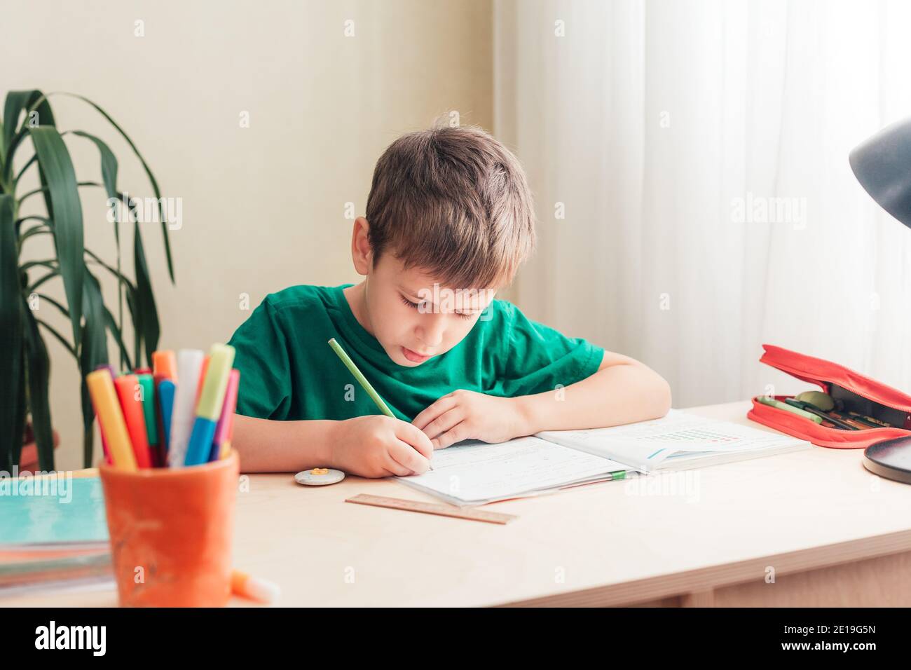 Intelligenter 7 Jahre alter Junge, der seine Schreibunterrichtstunde nach Hause macht, während er am Schreibtisch sitzt. Stockfoto
