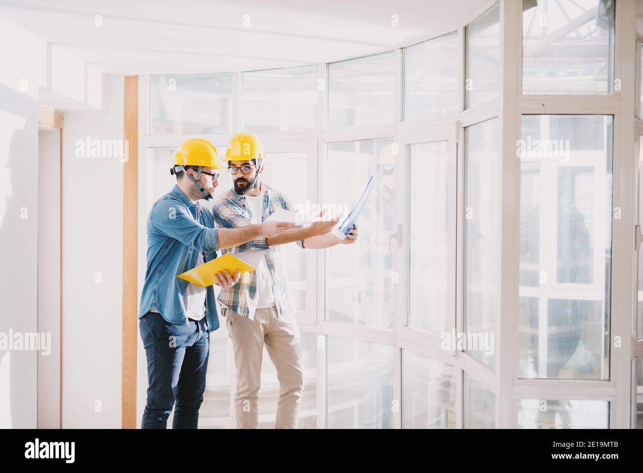 Zwei fokussierte junge professionelle Ingenieure mit Helmen, die im schönen großen hellen Büro mit großen Fenstern eine Diskussion über das Projekt führen. Stockfoto