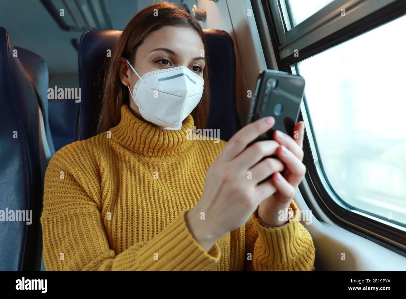 Entspannte Frau mit KN95 FFP2 Gesichtsmaske mit Smartphone-App. Zug Passagier mit Schutzmaske reisen in Business-Klasse SMS auf mobi sitzen Stockfoto