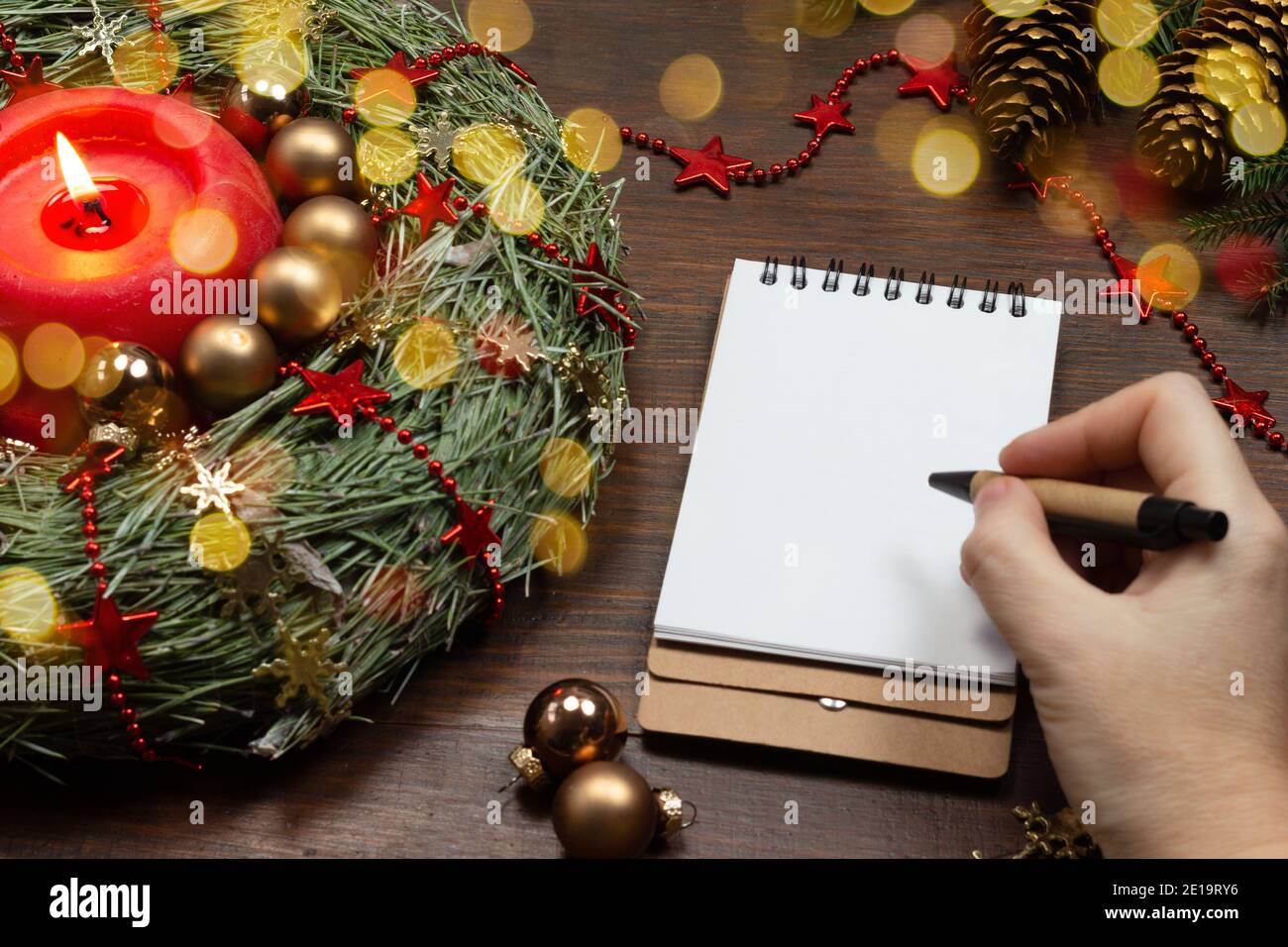 Eine Frauenhand hält einen Stift und schreibt in ein Notizbuch. Layout. Neujahrs Holztisch mit Tannenkranz, brennenden Kerzen und Weihnachtskugeln. Modell, bis Stockfoto