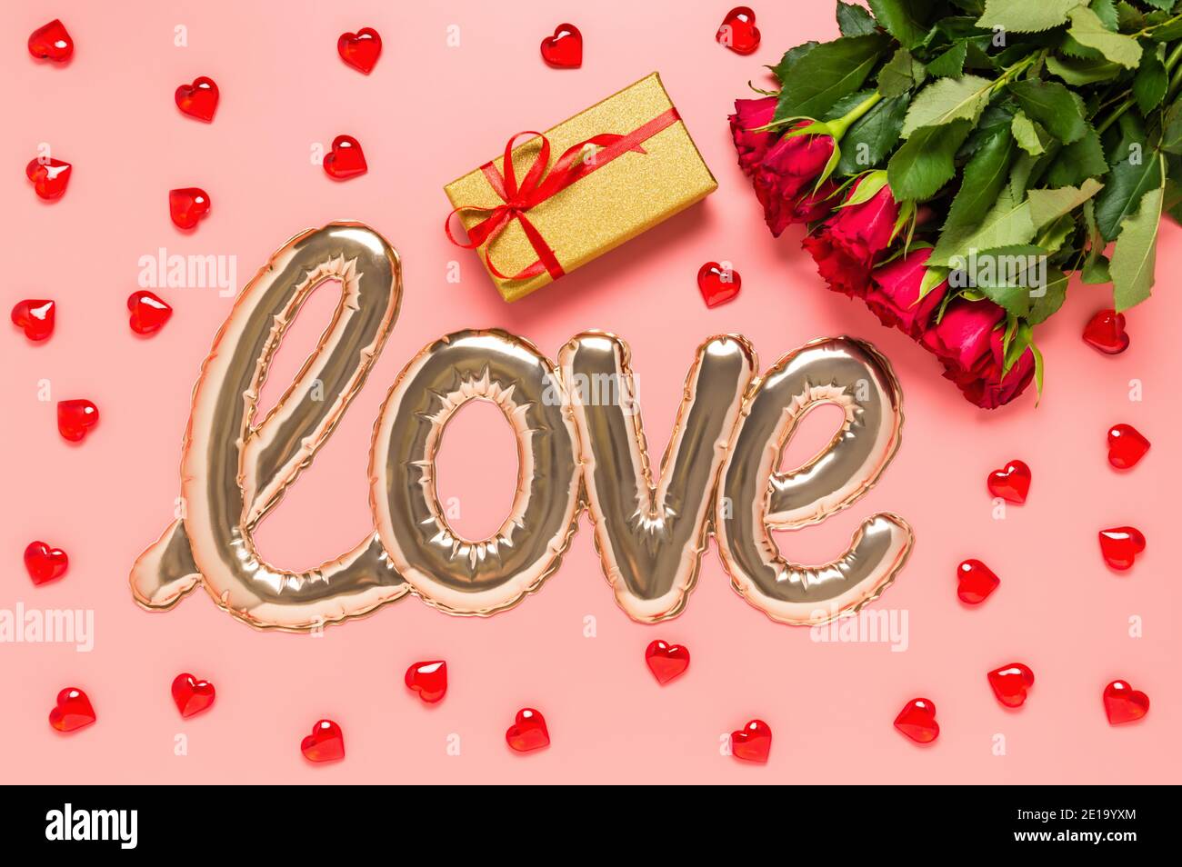 Liebeswort geformt mit glänzendem Folienballon, rotem Rosenstrauß, goldener Geschenkbox und Holzherzen auf rosa Hintergrund. Mütter, Frauen oder Valentinstag Stockfoto