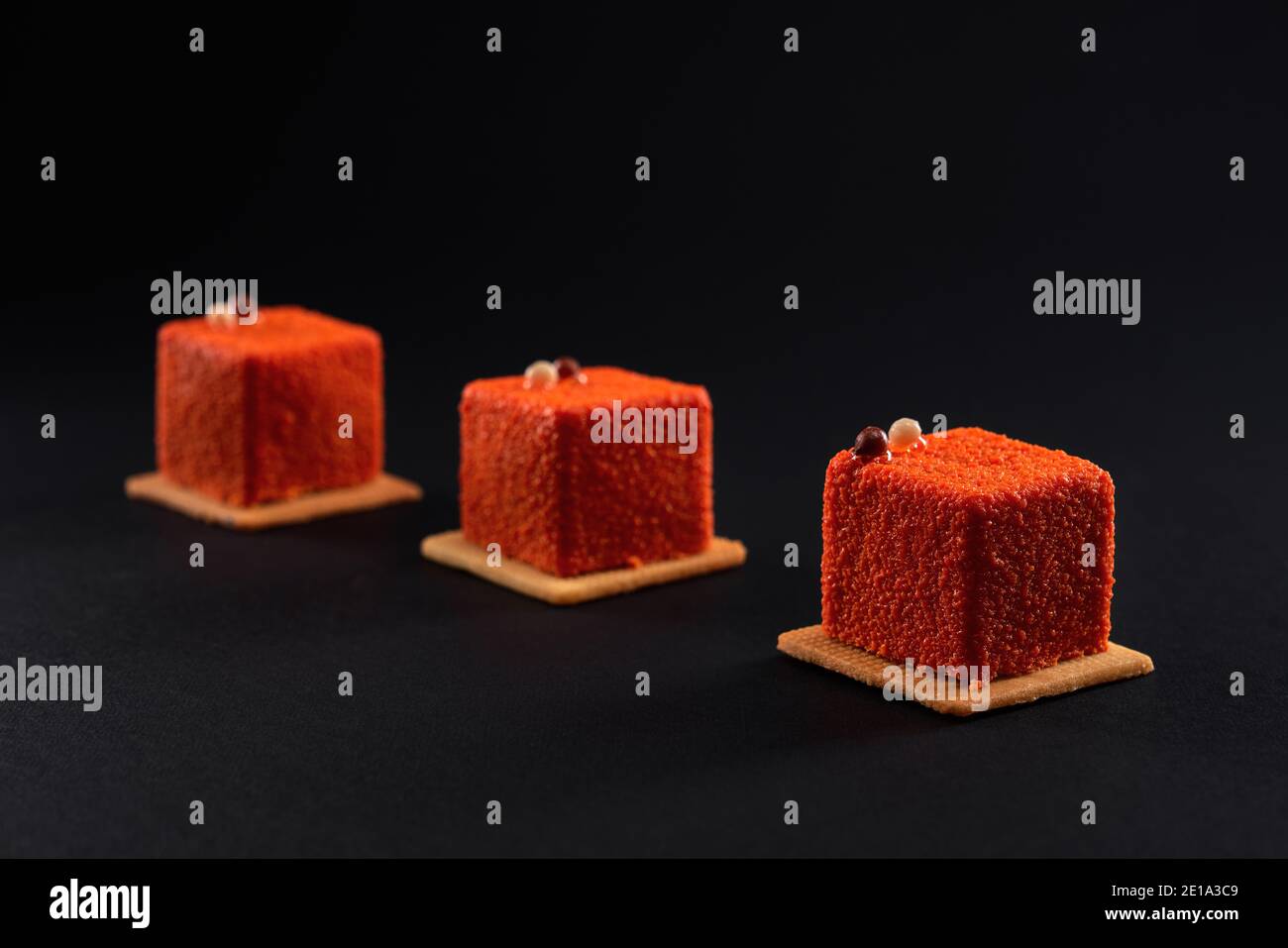 Nahaufnahme Ansicht von matt orange Dessert mit Schokoladenkugeln auf schwarzem Studio Hintergrund isoliert dekoriert. Drei kleine quadratische matte Kuchen in Reihe auf Cookies im Restaurant. Konzept der Süßwaren. Stockfoto