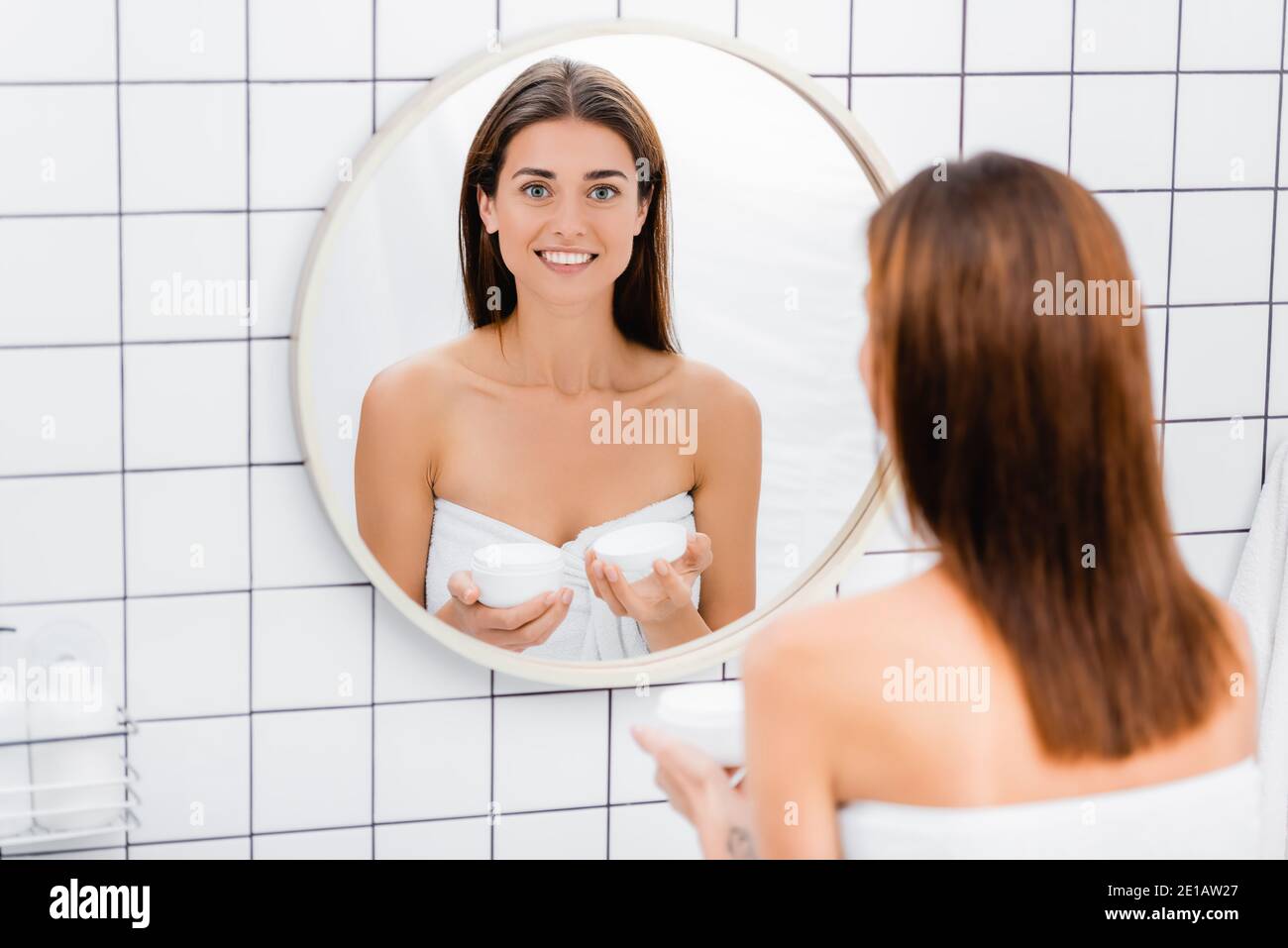 Glückliche junge Frau hält kosmetische Creme in der Nähe Spiegel im Badezimmer, verschwommen Vordergrund Stockfoto