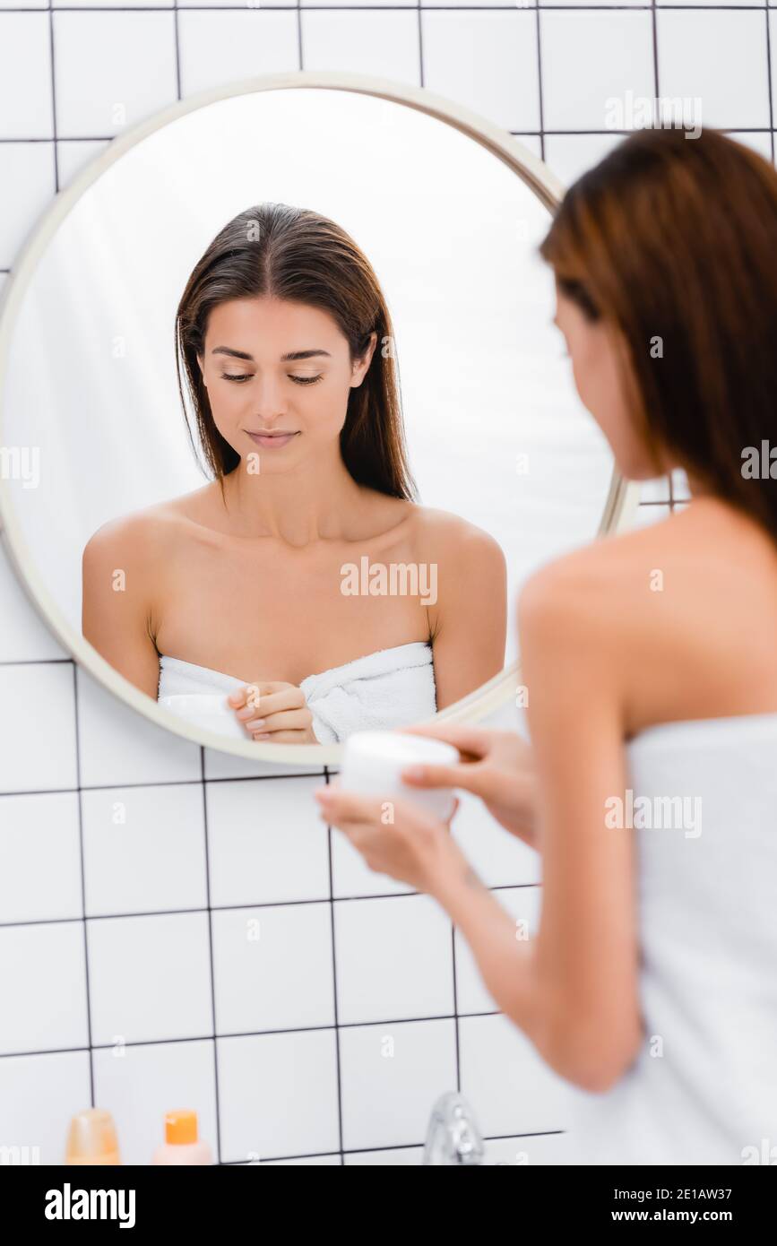 Junge Frau, in weißes Handtuch gewickelt, öffnet kosmetische Creme in der Nähe des Spiegels, verschwommen Vordergrund Stockfoto
