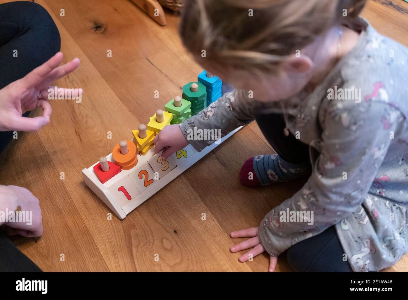 Eine Mutter, die ihre Finger hochhält, um ihrer zweijährigen Tochter zu zeigen, welche Nummer sie platzieren soll, indem sie ein Lernspielzeug mit Zählformen verwendet Stockfoto