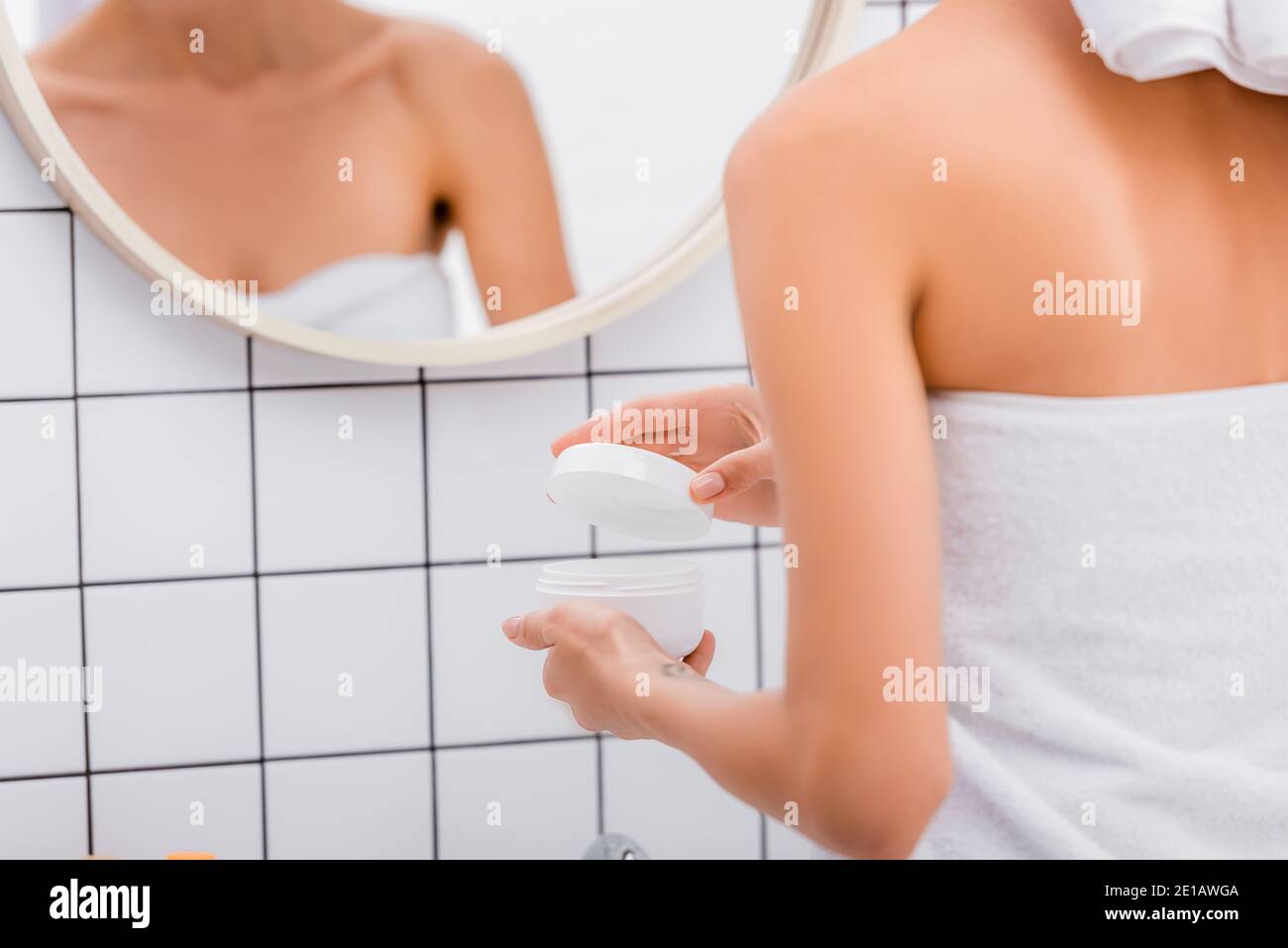 Beschnittene Ansicht der Frau, die kosmetische Creme in der Nähe verschwommener Reflexion öffnet Im Spiegel Stockfoto