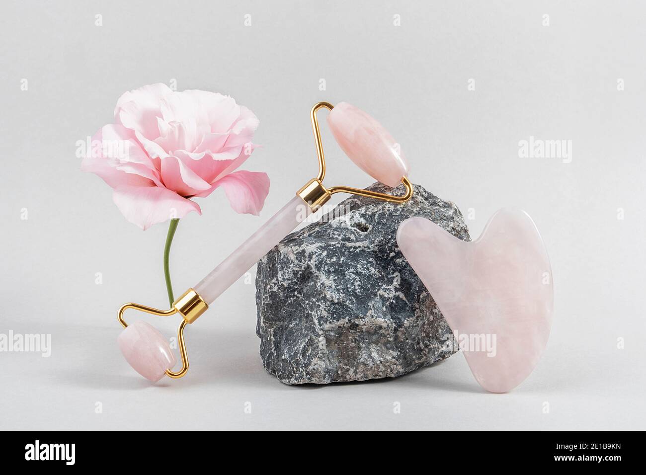 Crystal Rose Quarz Gesichtswalze und Massage-Tool Jade Gua Sha auf Steinen und rosa Blume auf grauem Hintergrund. Anti-Age-Gesichtsmassage für natürlichen Lift Stockfoto