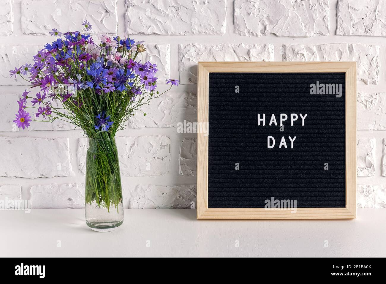 Happy Day Text auf schwarzem Briefbrett und Blumenstrauß blaue Blumen in Vase auf dem Tisch gegen weiße Ziegelwand. Stockfoto