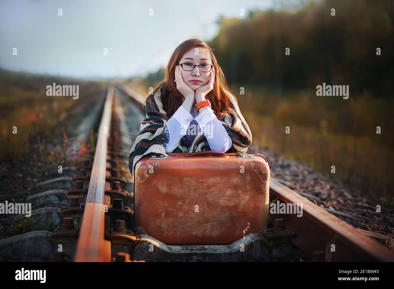 Eine schöne junge Frau in Brille und mit einem alten braunen Koffer sitzt auf den Gleisen und wartet leider auf die Ankunft des Zuges, um irgendwo hinzufahren. Jou Stockfoto
