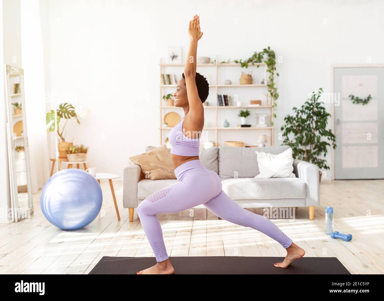 Seitenansicht einer jungen schwarzen Frau, die zu Hause Yoga praktiziert, in Kriegerpose steht und ihre Kraft trainiert Stockfoto