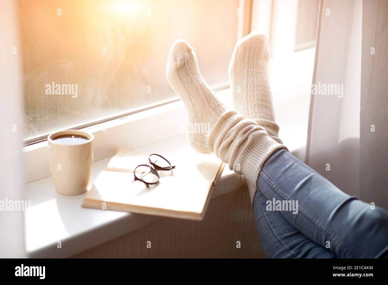 Beschnittene Ansicht der Frau, die ihre Füße in warme bequeme Socken auf Fensterbank mit Buch und Kaffee, Nahaufnahme Stockfoto