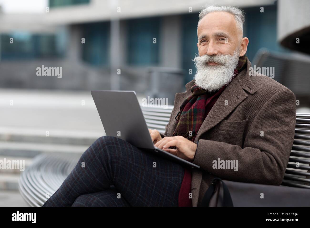 Wohlhabender grauhaariger Mann, der am Bürogebäude sitzt und einen Laptop benutzt Stockfoto