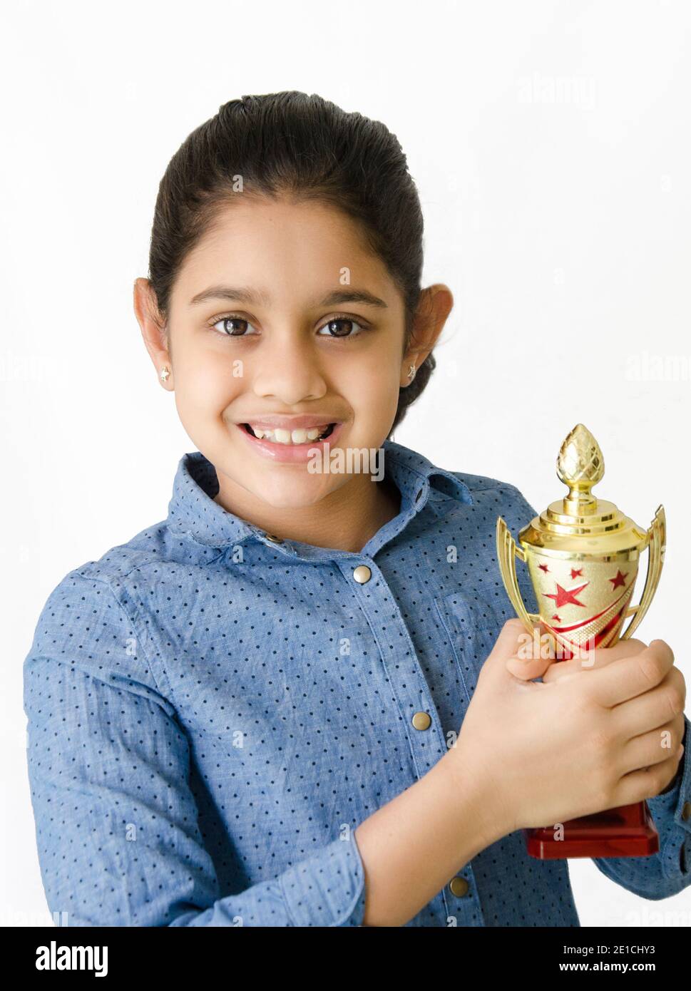 Portrait von einem schönen und lächelnden indischen Schulmädchen in weißem Hintergrund mit einer goldenen Trophäe und Darstellung Sieg. Stockfoto