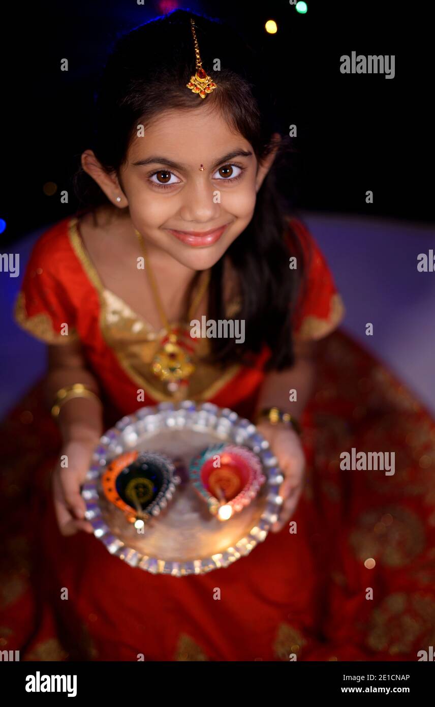 Ein schönes lächelndes indisches Mädchen in ethnischer Kleidung mit Juwelen gekleidet und hält Öllampen feiern diwali in der Nacht mit schwachen Lichtern im Hintergrund. Stockfoto