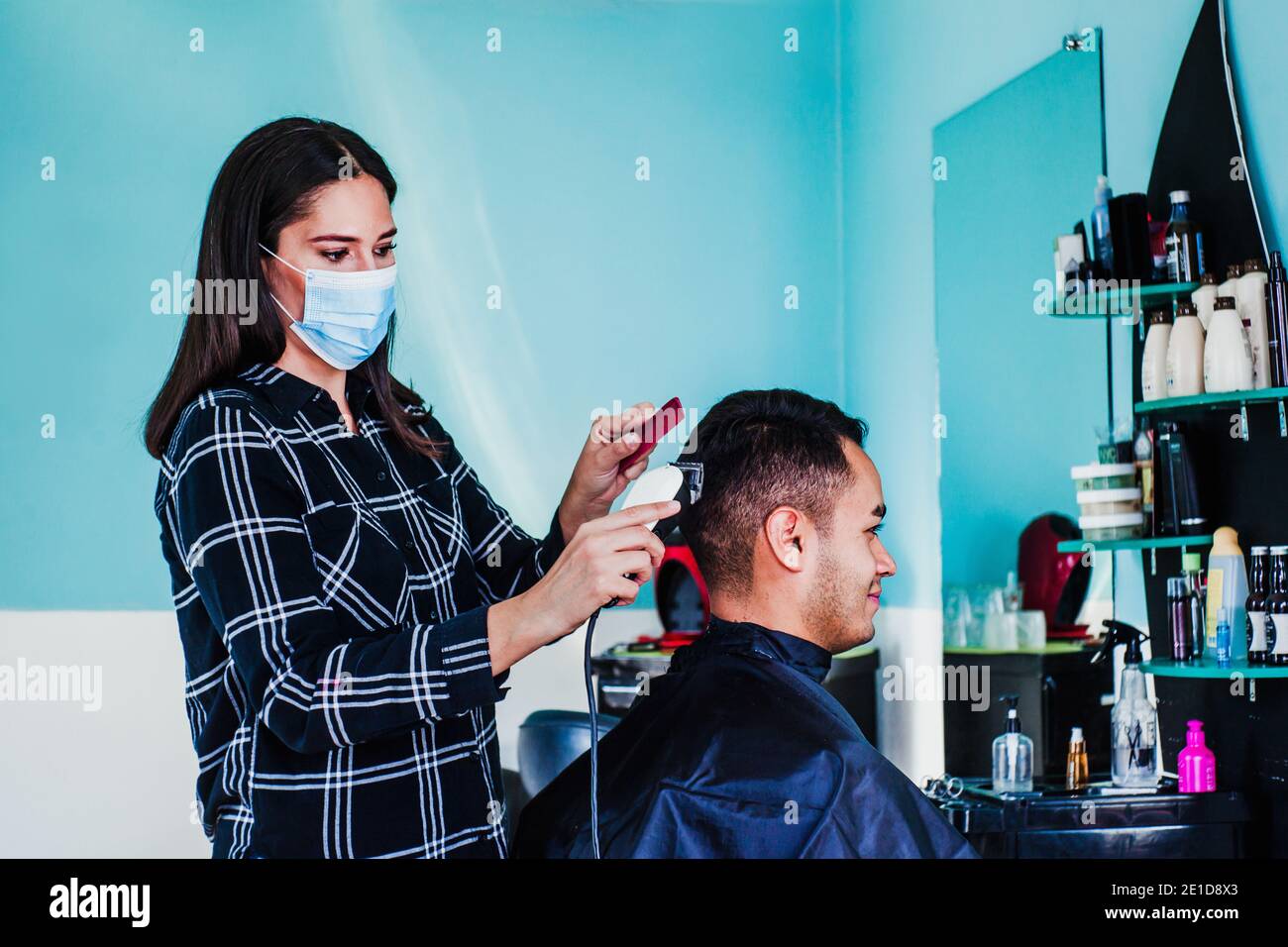 latin Frau Friseur mit Gesichtsmaske Schneiden Haare mit Haarschneidemaschine und Kamm. Mexikanischer Mann bekommt einen Haarschnitt. Barbier-Service mit Sicherheitsmaßnahmen Stockfoto