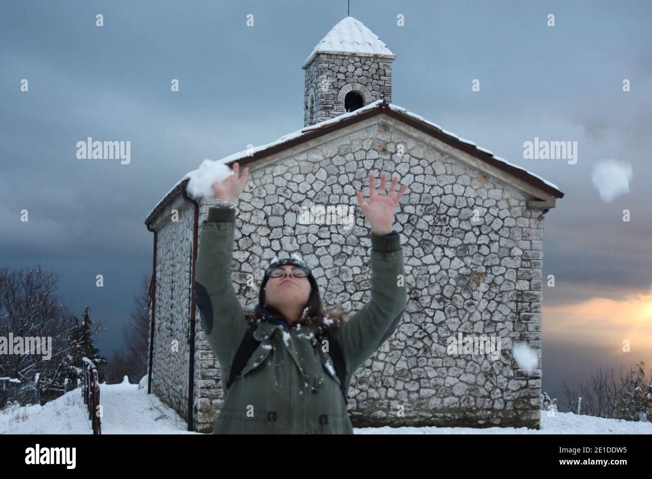 Mädchen im Winter Overalls spielt mit weichem weißen Schnee in Eine wunderschöne Sonnenuntergangslandschaft am Meer unter einer Kirche Mit Kirchturm in den Bergen in apuan al Stockfoto