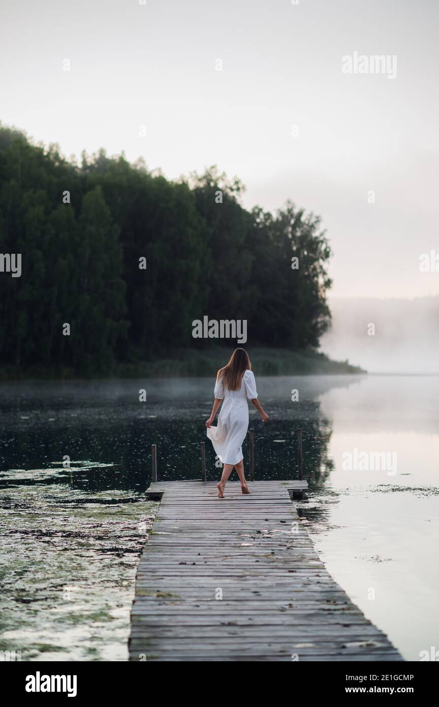Junge Frau in weißem Kleid auf einem See an einem kühlen Morgen mit einem Nebel über Wasser. Stockfoto