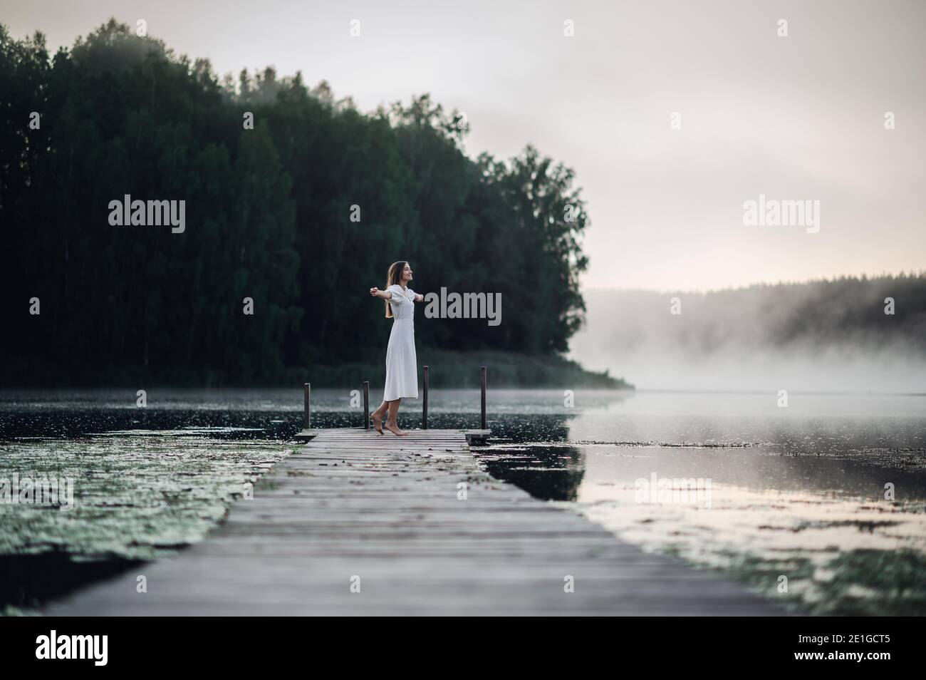 Glückliche Frau in einem weißen Kleid genießt den Morgen auf einem See. Nebel schwimmt über dem Wasser. Stockfoto