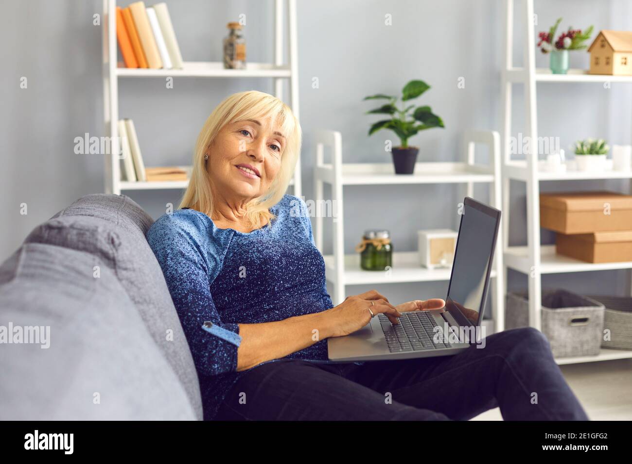 Lächelnde blonde Frau mittleren Alters, die auf dem Sofa sitzt und arbeitet Online auf dem Laptop zu Hause Stockfoto