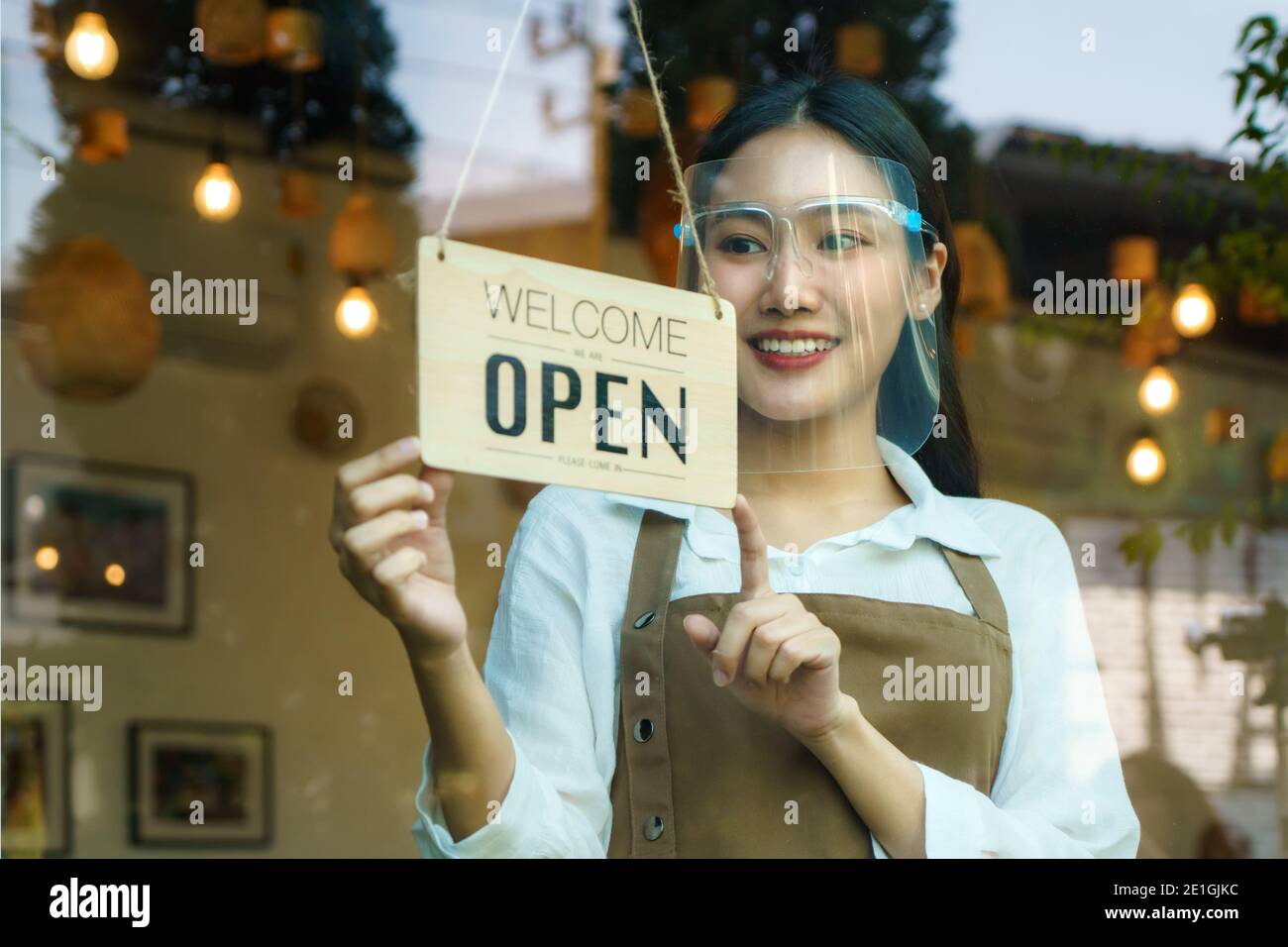 Asiatische Geschäftseigentümer Frau tragen Schutzschild hängend offen Schild in ihrem Restaurant / Café, wieder geöffnet nach Lockdown für neue normale Lebensweise Stockfoto