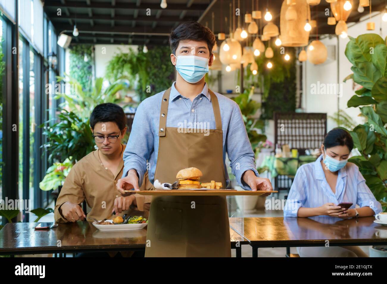 Portrait attraktive asiatische Kellnerin tragen Gesichtsmaske und Gesichtsschutz halten Lebensmittel Tablett zu servieren Mahlzeit an Kunden mit custome im Hintergrund. Neue normale Stockfoto