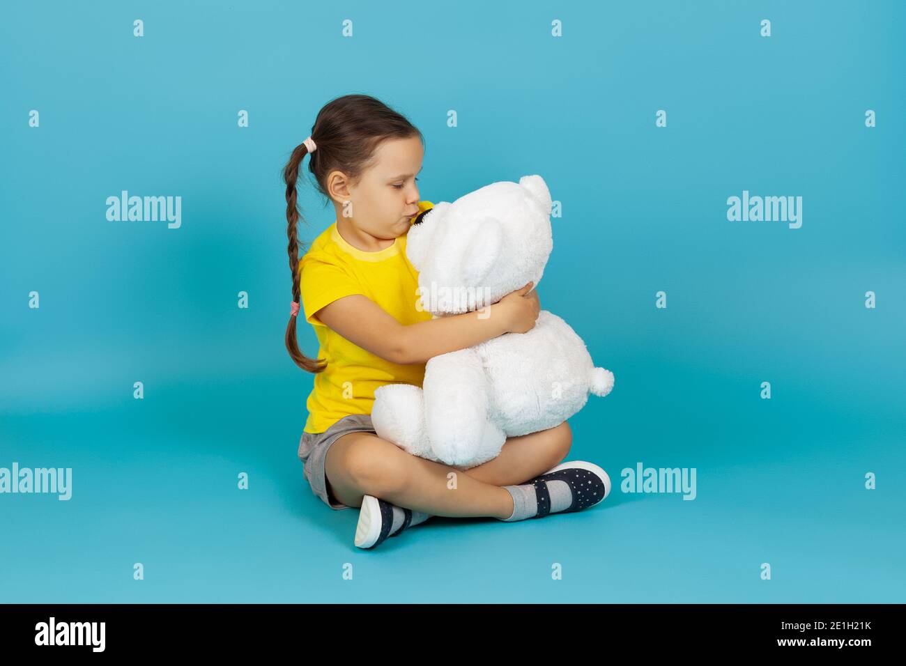 Niedliches Kind mit Zöpfen sitzt mit gekreuzten Beinen auf dem Boden, umarmt einen weißen Teddybären und küsst seine Nase isoliert auf einem blauen Hintergrund Stockfoto